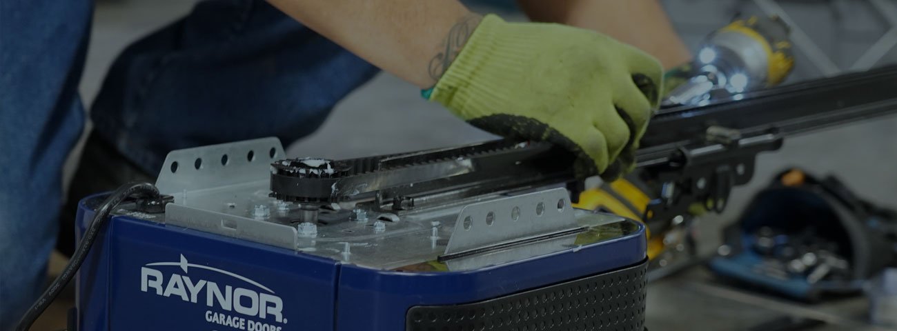 A technician in work gloves servicing the motor unit of a blue Raynor garage door opener