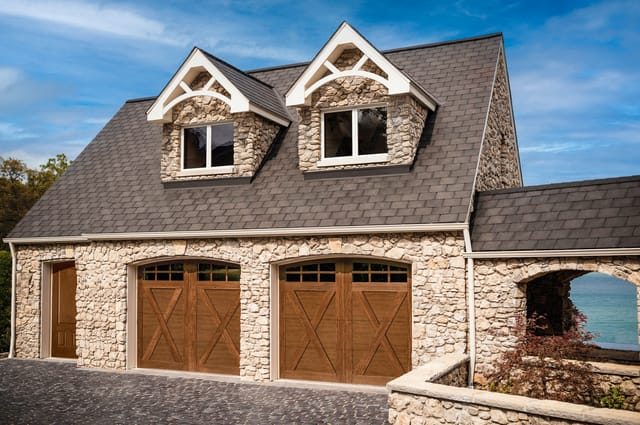 Clopay Carriage House dark wooden garage doors on stone-clad house with dormer windows.