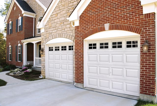 Clopay Classic Steel Gold-Series white colonial garage doors on adjacent brick and stone houses with concrete driveway.
