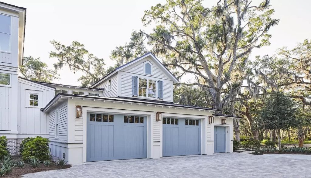 Exterior view of white house with blue CloPay garage doors, dormer window, outdoor lanterns, large moss-draped tree, and paved driveway.