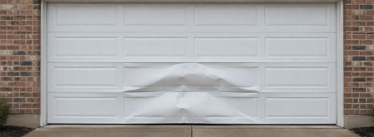 A white garage door with its bottom two panels creased and pushed inward, framed by brick walls on a concrete driveway