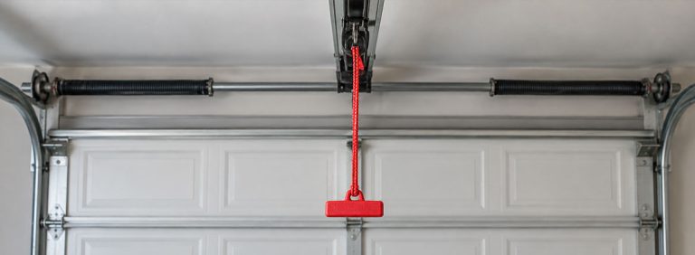 A red emergency release cord with a red handle hanging from the opener trolley above a fully closed white garage door, with the torsion springs on the bar overhead